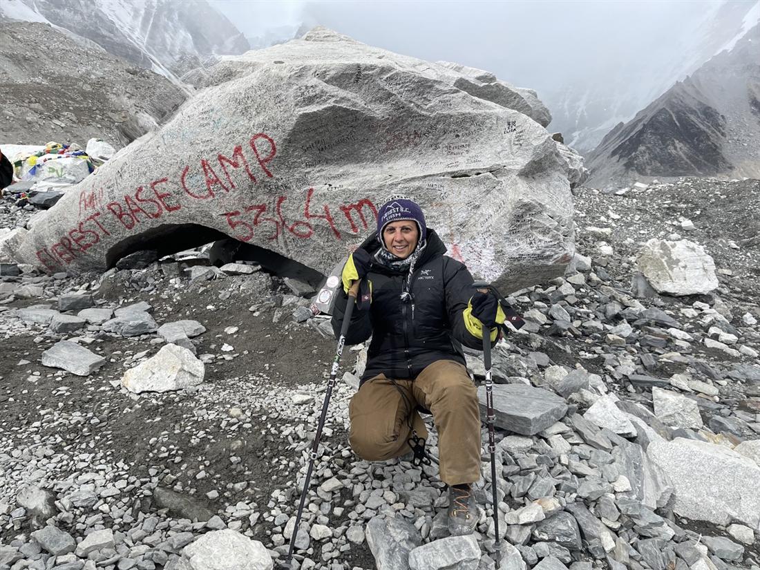 AnnMarie Castrigno kneeling with trekking poles at the Everest Base Camp marker stone, painted with Everest Base Camp 5364m